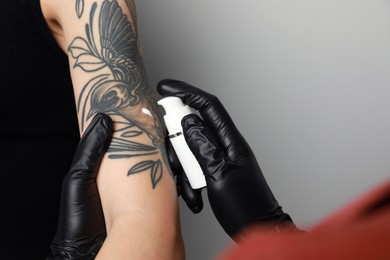 Worker in gloves applying cream on woman's arm with tattoo against light background, closeup