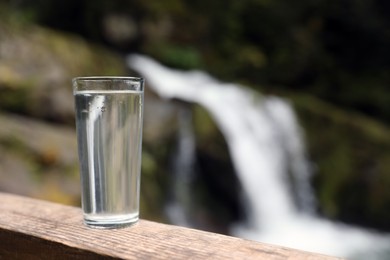 Photo of Glass of water on wooden surface near waterfall outdoors. Space for text