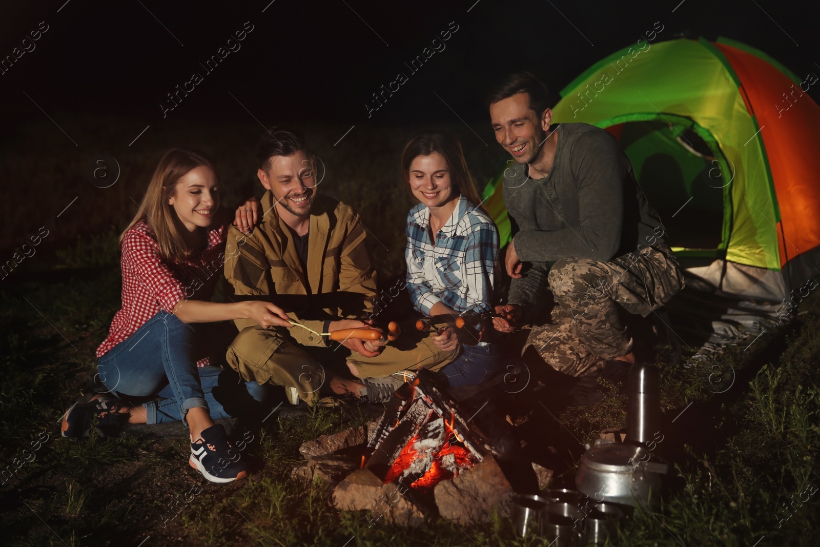Photo of Friends frying sausages on bonfire at night. Camping season