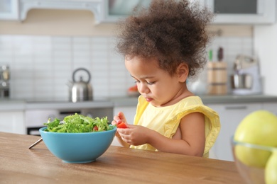 Photo of Cute African-American girl eating vegetable salad at table in kitchen