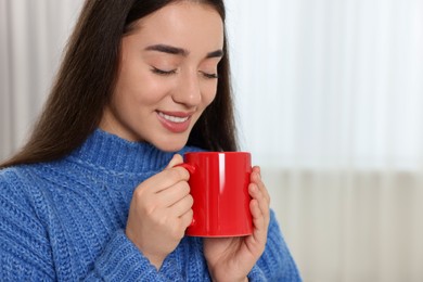 Photo of Happy young woman holding red ceramic mug at home, space for text