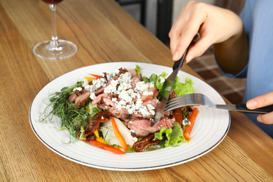 Photo of Woman eating delicious salad with roasted meat at wooden table, closeup