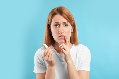 Upset woman with herpes applying cream on lips against light blue background