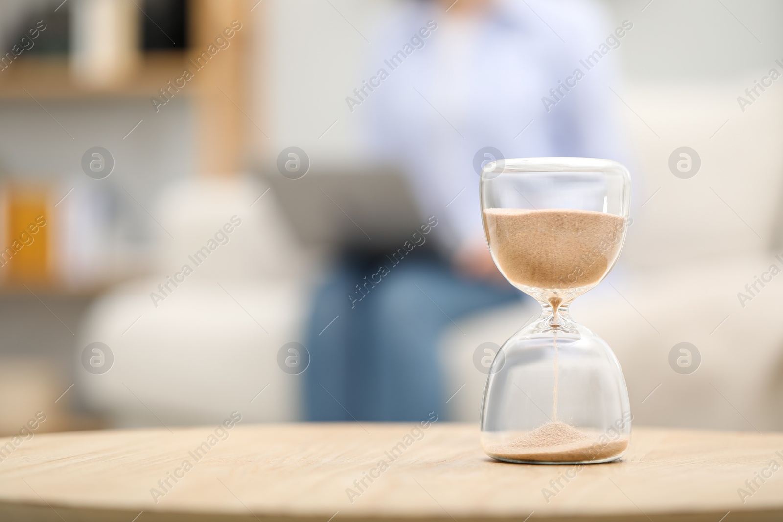 Photo of Hourglass with flowing sand on desk. Woman using laptop indoors, selective focus