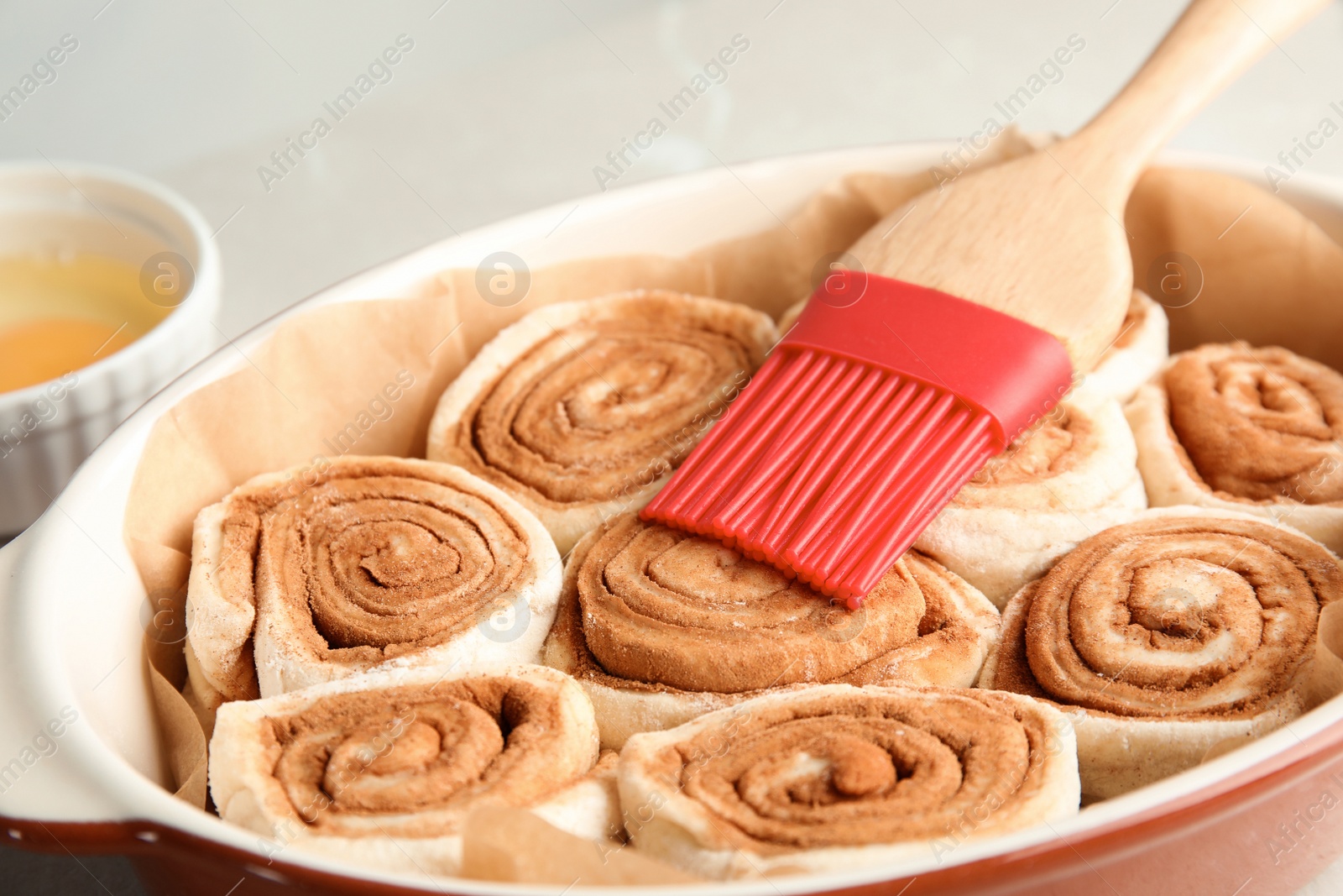 Photo of Baking dish with raw cinnamon rolls and pastry brush on table, closeup