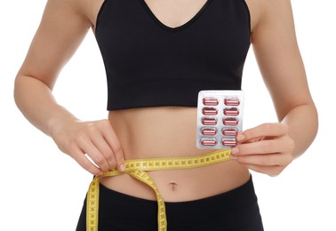 Photo of Slim woman holding pills and measuring waist with tape on white background, closeup. Weight loss
