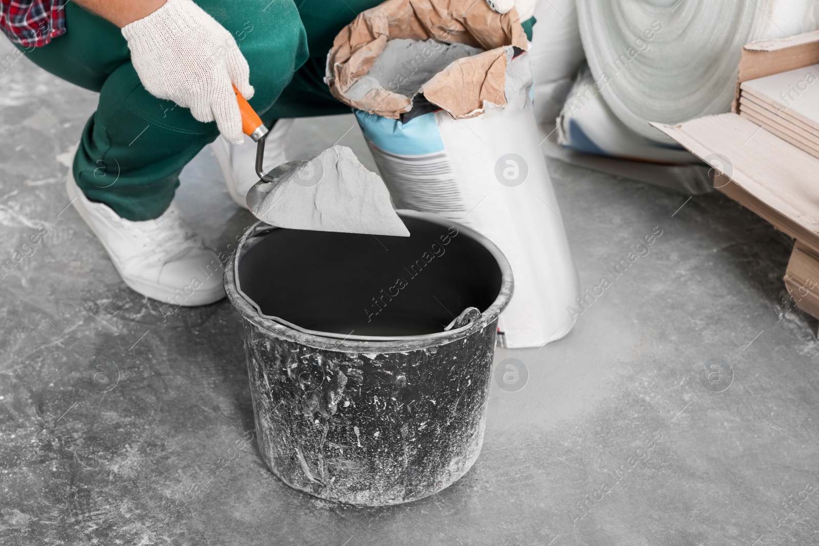 Photo of Worker with cement powder and trowel mixing concrete in bucket indoors, closeup