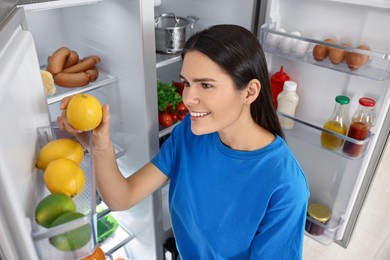 Photo of Young woman taking lemon out of refrigerator indoors, above view