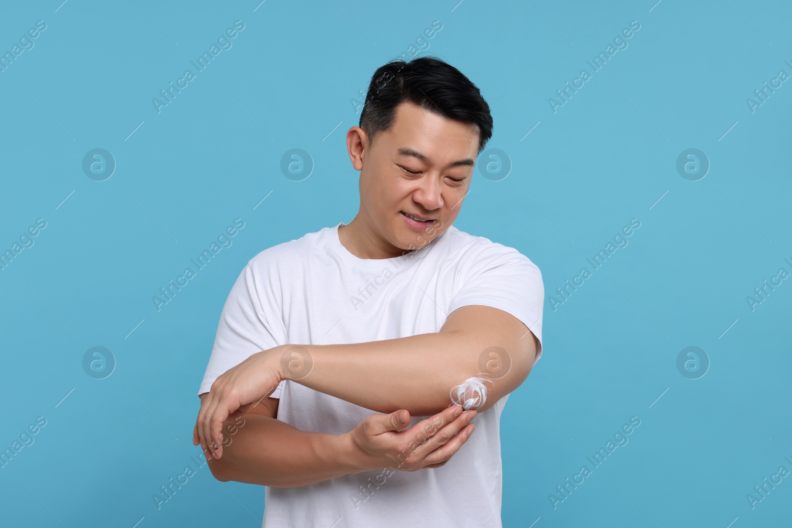 Photo of Handsome man applying body cream onto his elbow on light blue background