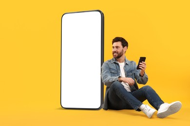 Image of Man with mobile phone sitting near huge device with empty screen on orange background. Mockup for design