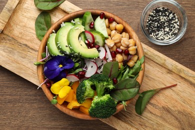 Delicious vegan bowl with broccoli, avocados and violet flowers on wooden table, flat lay