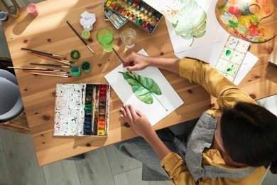 Young woman drawing leaf with watercolors at table indoors, top view