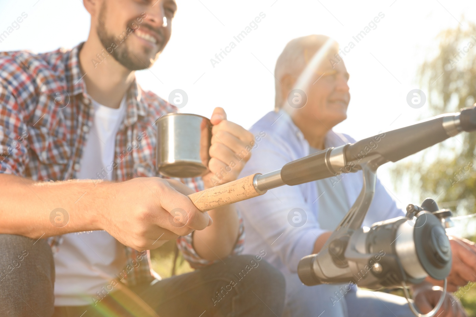 Photo of Father and adult son fishing together from riverside on sunny day