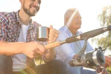 Father and adult son fishing together from riverside on sunny day