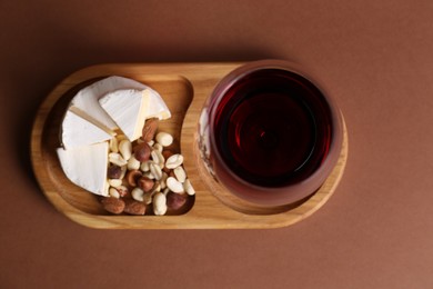 Photo of Glass of red wine, cheese and nuts on brown background, top view
