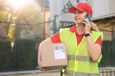 Photo of Courier in uniform with parcel talking on smartphone near private house outdoors, space for text