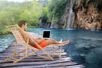 Image of Young man with laptop relaxing on sun lounger near lake and waterfall. Luxury vacation 