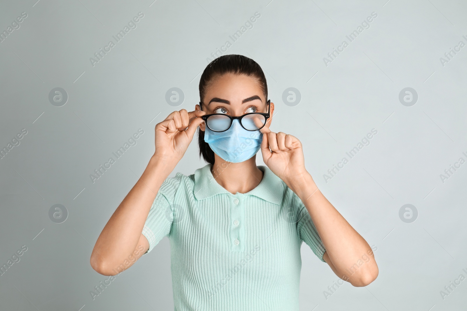 Photo of Woman wiping foggy glasses caused by wearing medical mask on light background