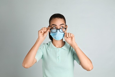 Photo of Woman wiping foggy glasses caused by wearing medical mask on light background