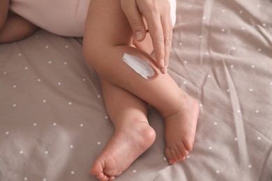 Photo of Mother applying moisturizing cream onto her little baby's skin on bed, closeup