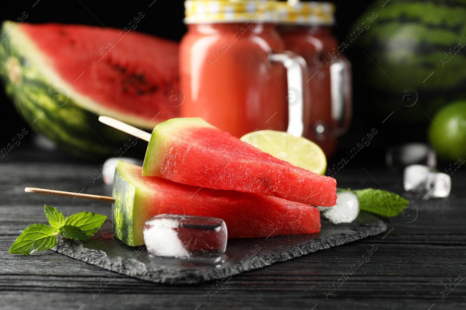 Photo of Slate board with juicy watermelon, ice and lime on black wooden table, closeup