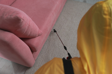 Photo of Pest control worker in protective suit spraying insecticide near sofa indoors, above view
