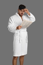 Young man in bathrobe reading newspaper on grey background