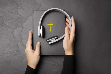 Woman with Bible and headphones at black table, top view. Religious audiobook