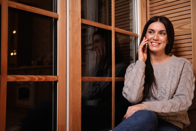 Beautiful woman talking on mobile phone near window indoors. Christmas celebration