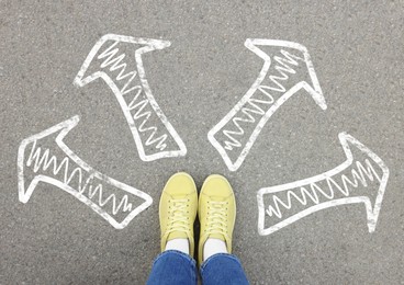 Choosing future profession. Girl standing in front of drawn signs on asphalt, top view. Arrows pointing in different directions symbolizing diversity of opportunities