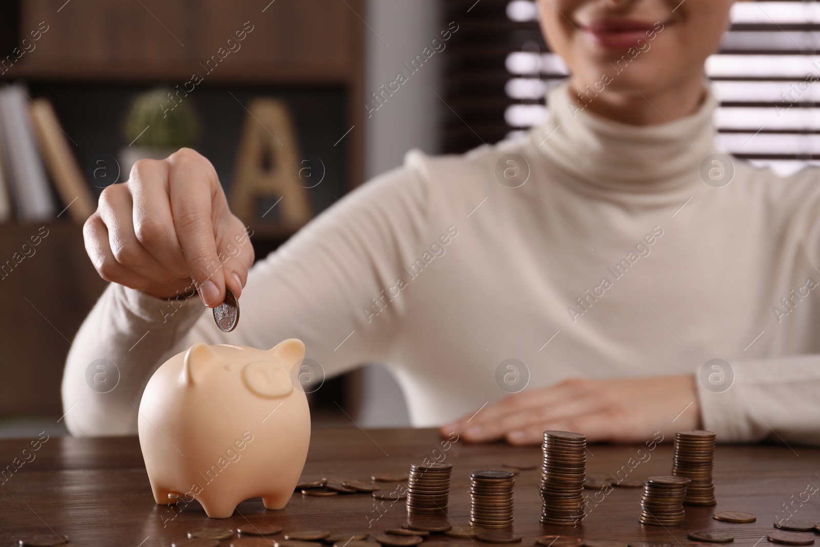 Photo of Woman putting coin into piggy bank at wooden table indoors, closeup