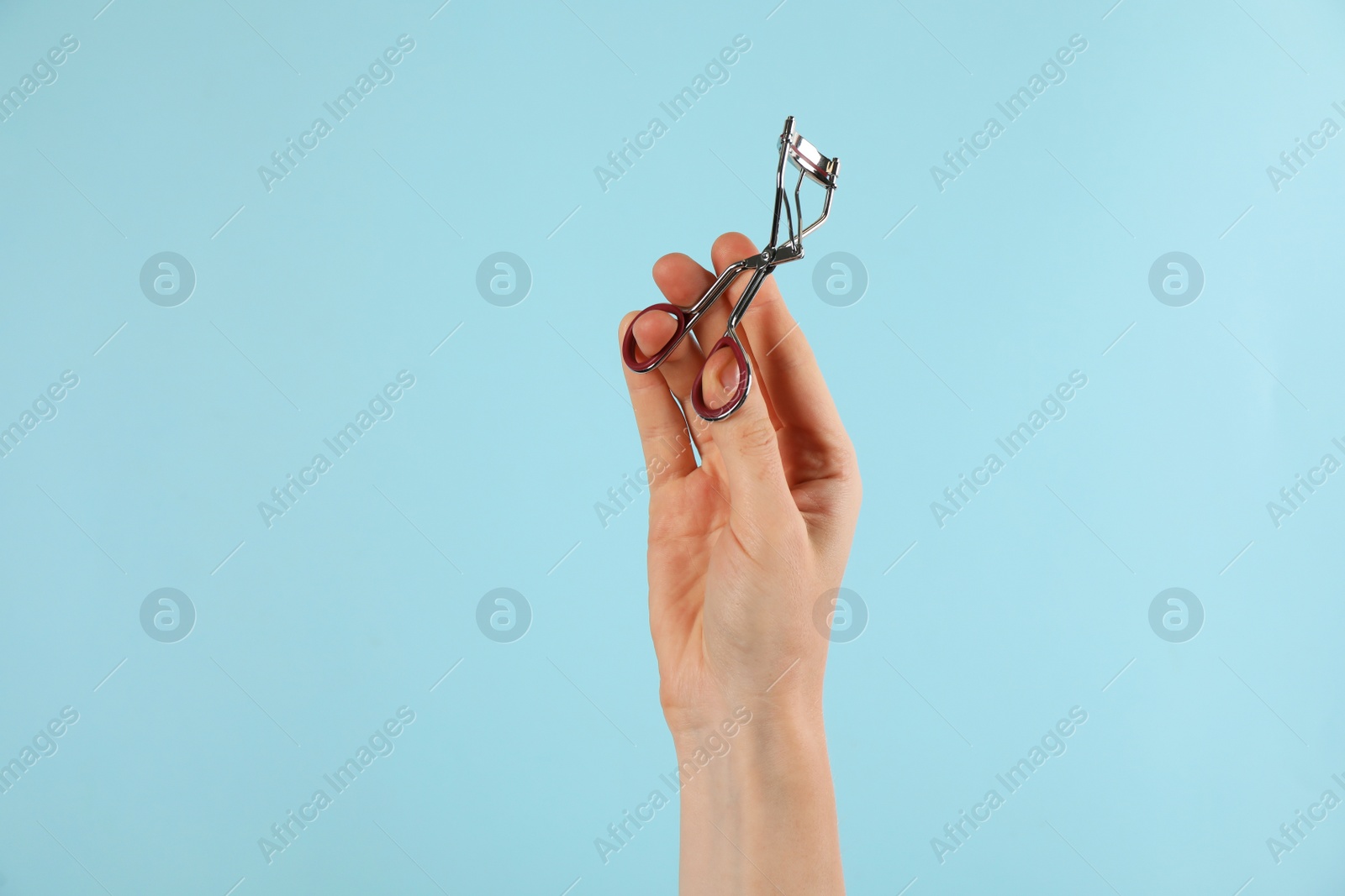 Photo of Woman holding eyelash curler on light blue background, closeup. Makeup tool