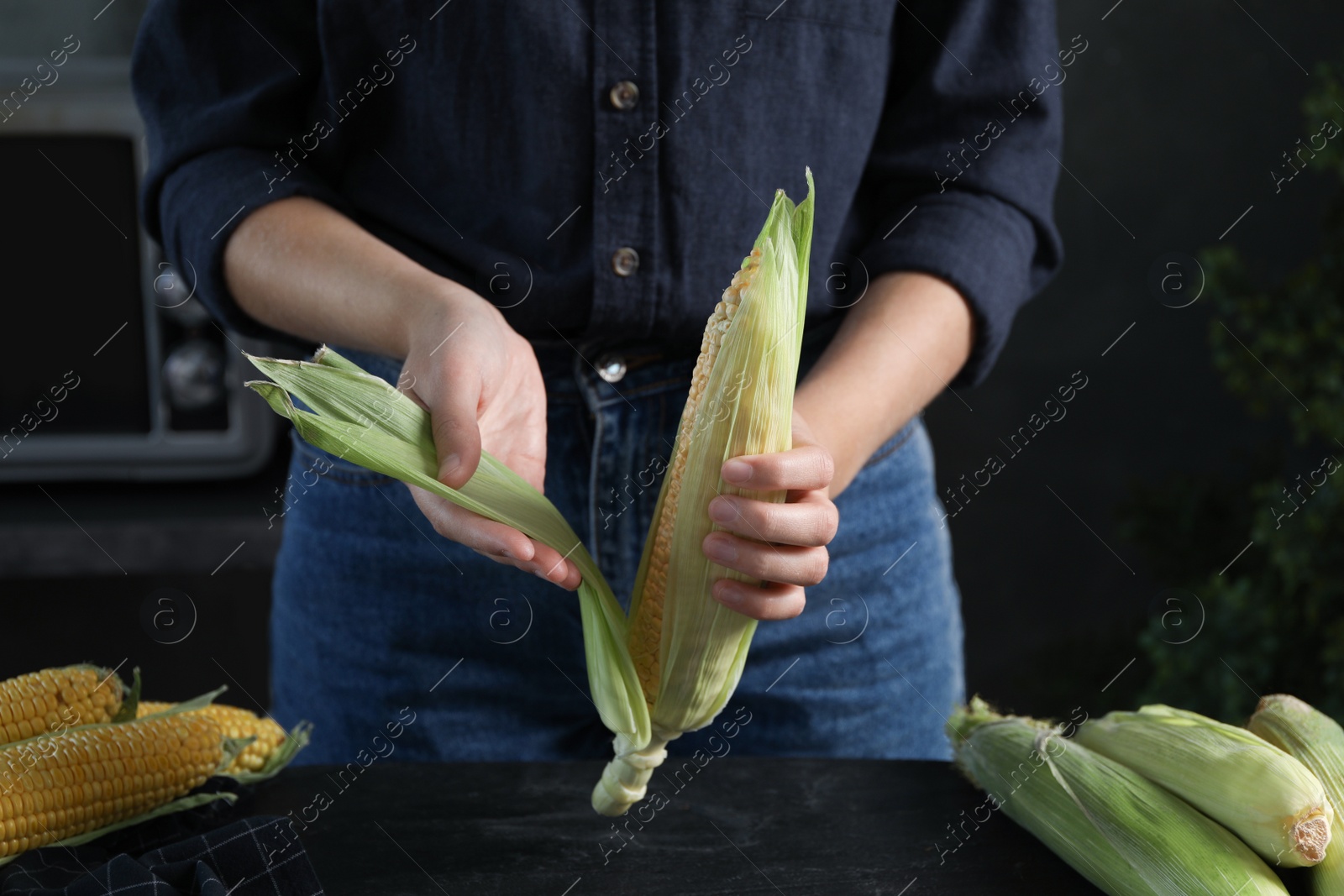 Photo of Woman husking corn cob at black table, closeup