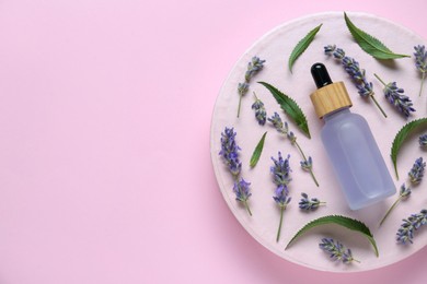 Bottle of lavender essential oil surrounded by leaves and flowers on pink background, top view. Space for text