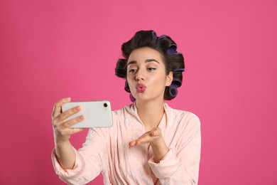 Photo of Young woman in bathrobe with hair curlers taking selfie on pink background