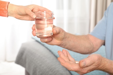 Photo of Woman giving glass of water to senior man with pill indoors, closeup