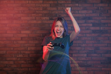 Emotional young woman playing video games with controller near brick wall