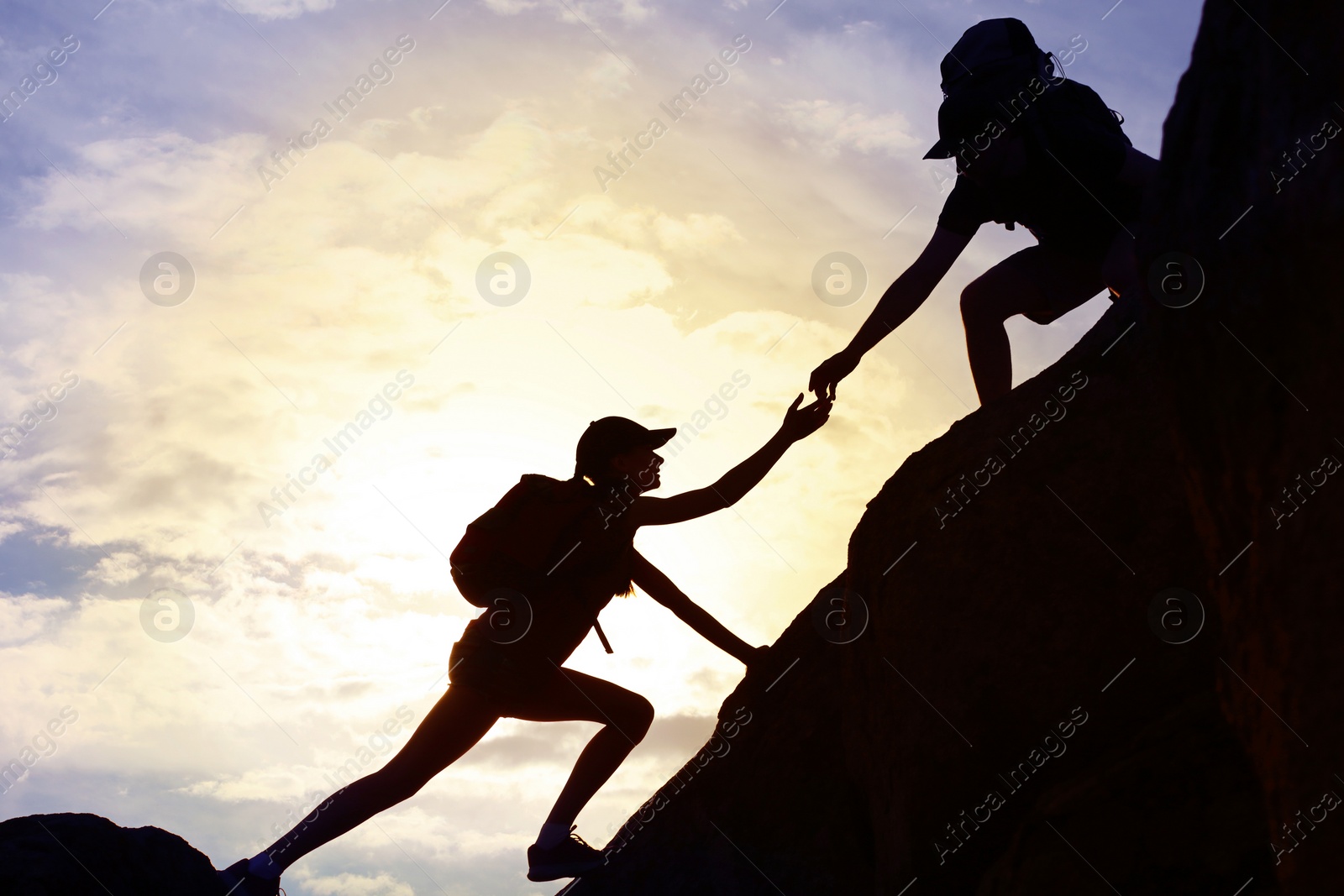 Photo of Silhouettes of man and woman helping each other to climb on hill against sunset