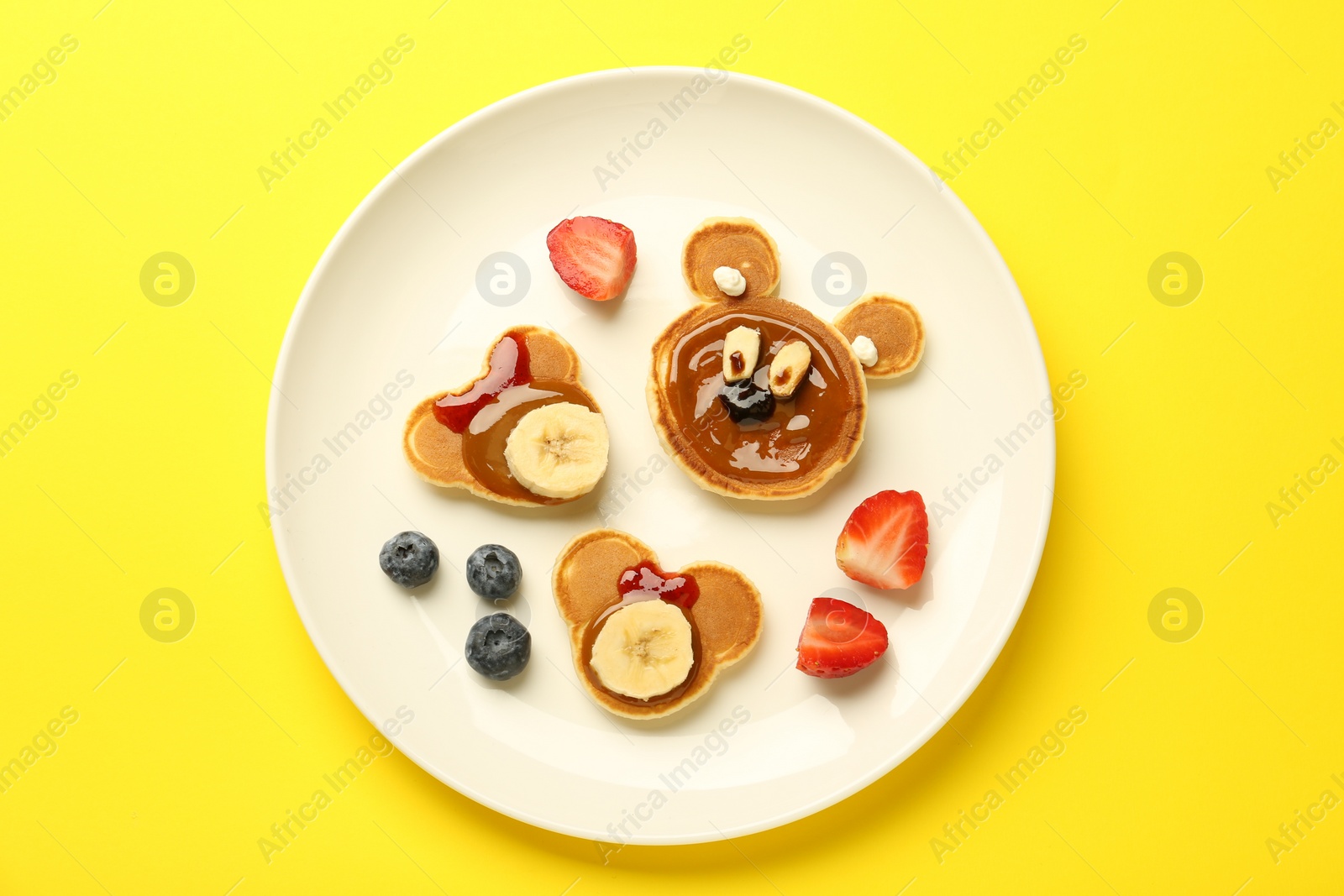 Photo of Creative serving for kids. Plate with cute bears made of pancakes, berries, banana and chocolate paste on yellow background, top view