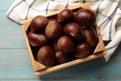 Roasted edible sweet chestnuts in crate on light blue wooden table, top view