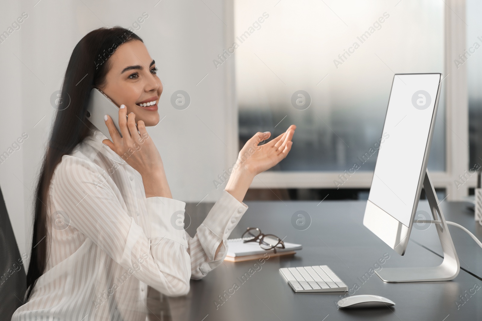 Photo of Happy woman using modern computer while talking on smartphone at black desk in office