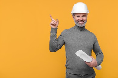 Photo of Architect in hard hat holding draft and pointing at something on orange background. Space for text