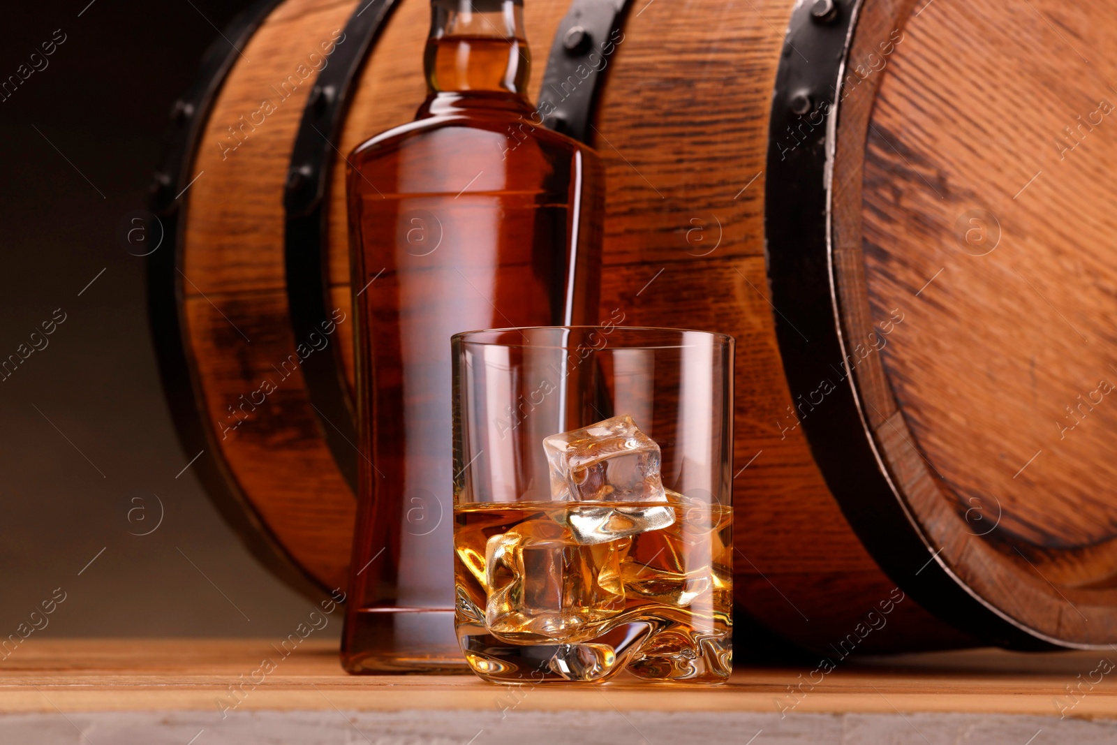 Photo of Whiskey with ice cubes in glass, bottle and barrel on wooden table against dark background, closeup