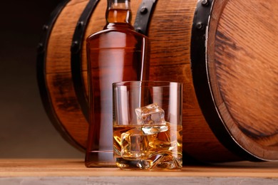 Whiskey with ice cubes in glass, bottle and barrel on wooden table against dark background, closeup