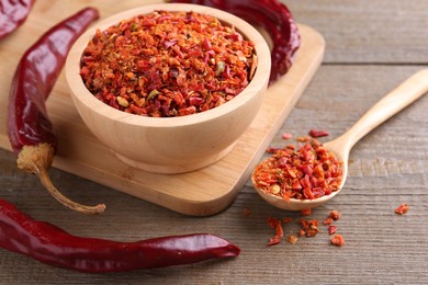 Photo of Aromatic spice. Red chili pepper flakes in bowl, spoon and pods on wooden table, closeup