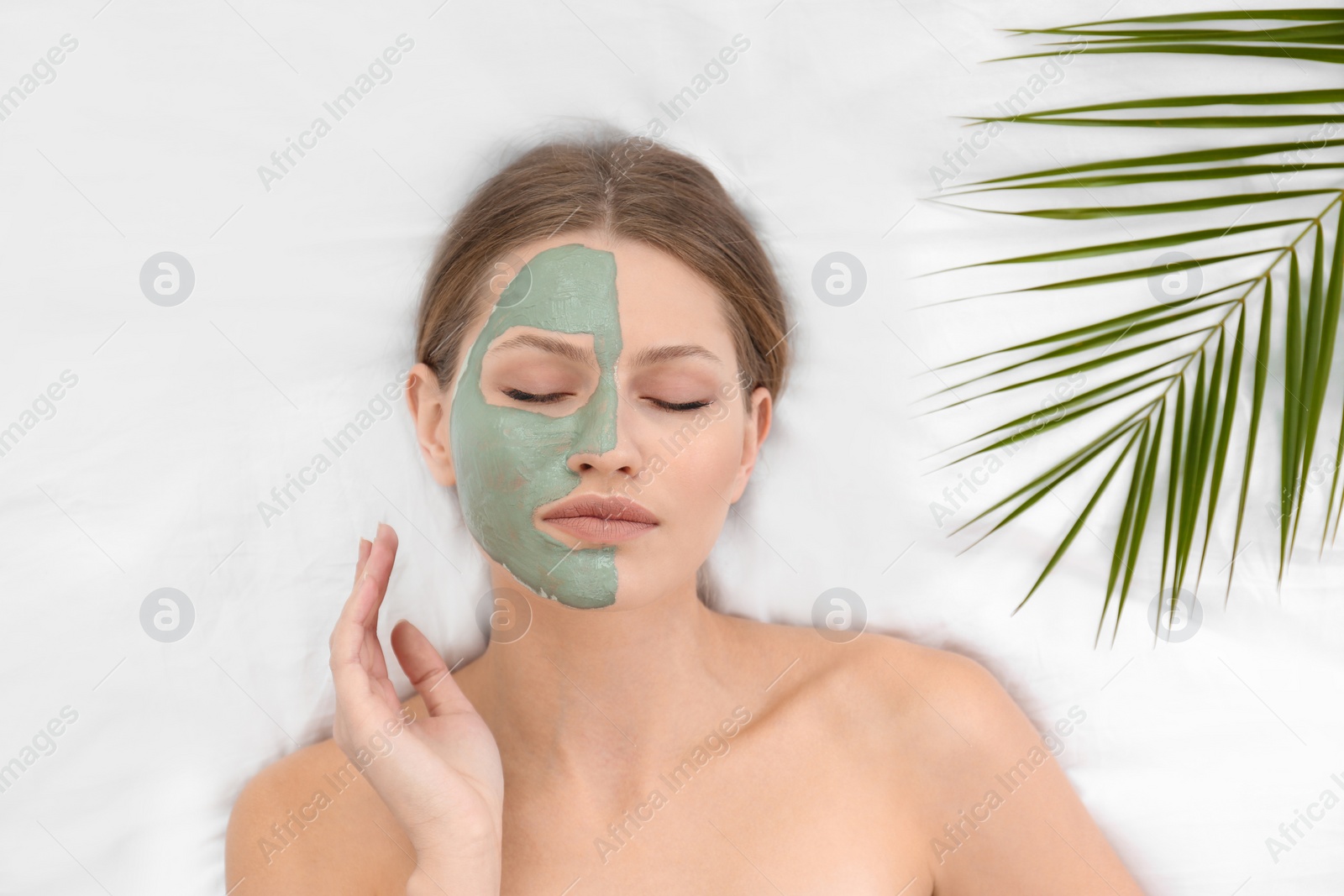 Photo of Beautiful woman with clay facial mask and tropical leaf on white fabric, above view