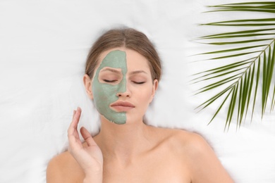 Beautiful woman with clay facial mask and tropical leaf on white fabric, above view