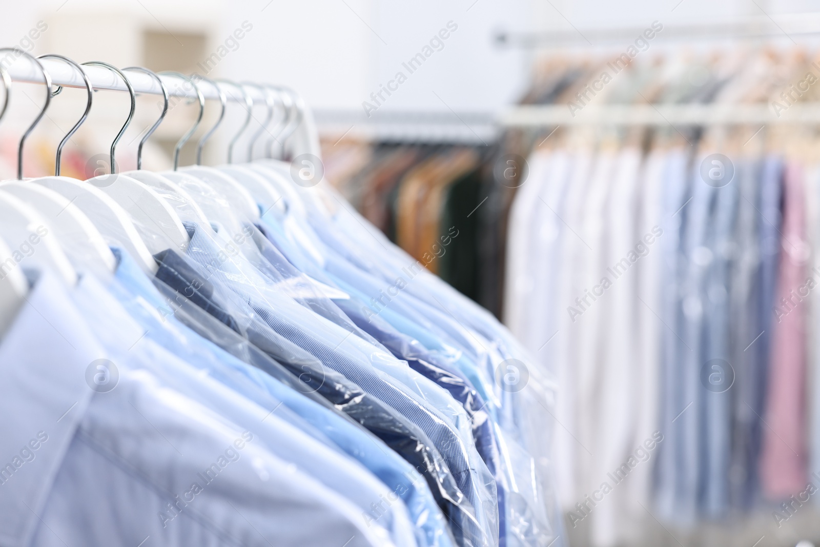 Photo of Dry-cleaning service. Many different clothes in plastic bags hanging on rack indoors, closeup