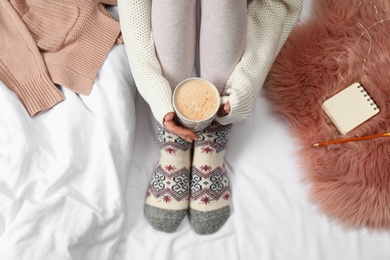 Woman with cup of hot drink wearing knitted socks on white fabric, top view. Warm clothes