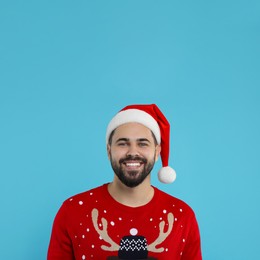 Photo of Happy young man in Christmas sweater and Santa hat on light blue background
